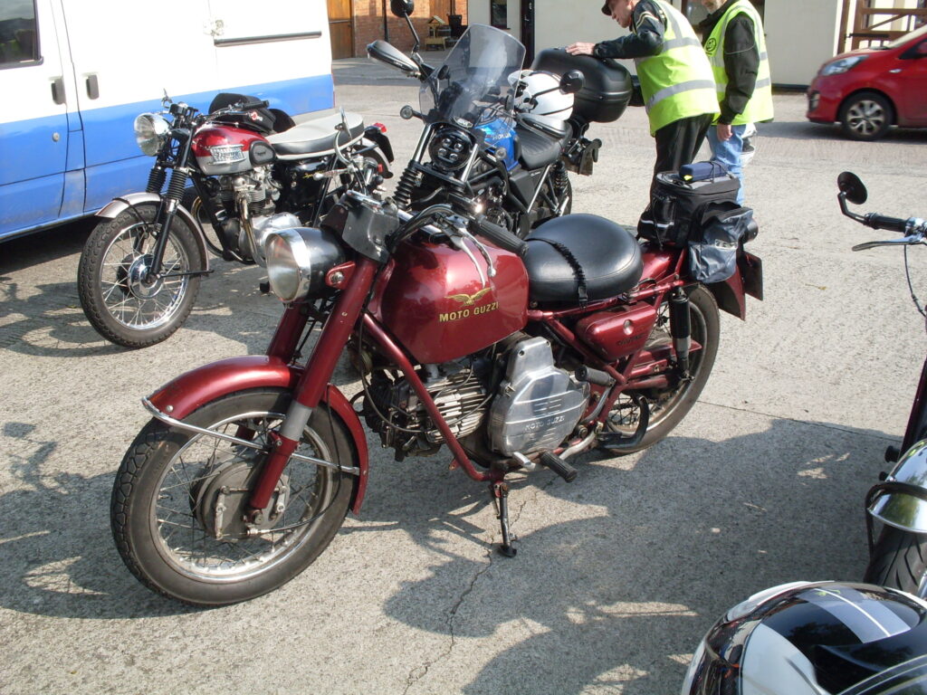 A red single (horizontal) cylinder Moto Guzzi of the type with a huge exposed flywheel next to the riders left leg only this model has the flywheel covered up...