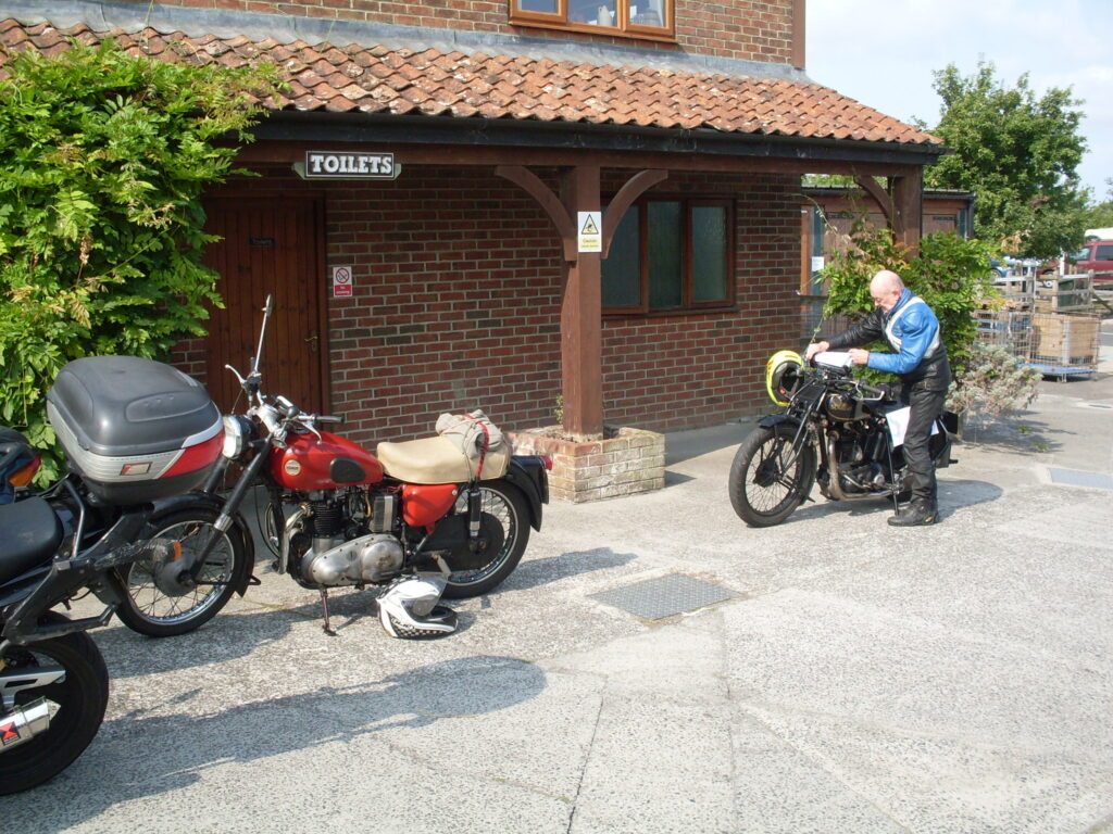 A red Ariel twin-cylinder motorbike from the 1950s and behind it a Sunbeam from the 1930s is having the paper route sheet loaded into the route-reader on the handlebars.
