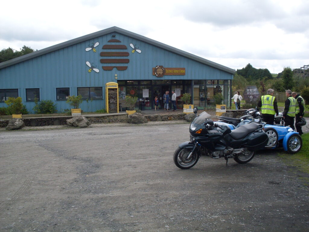 A small group of parked motorcycles and the background taken up with the low, wide, blue frontage of the 'Quince Honey Farm' building.