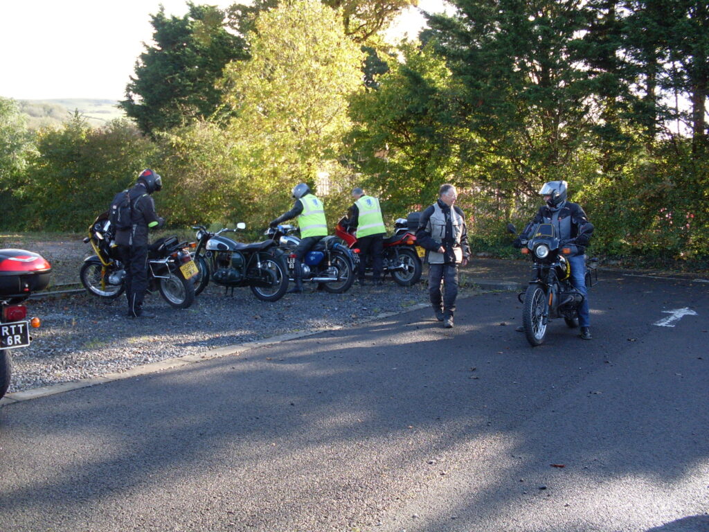 A small group of motorcyclists 'starting up' against a background of trees backlit by sunshine and with foliage just starting to 'turn' in autumn