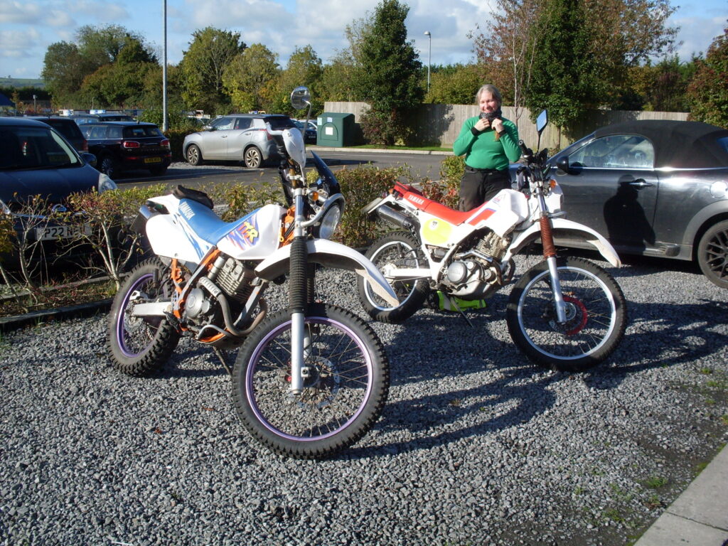 Two whites 'trail' bikes with knobbly tyres and lots of ground clearance, one female rider is pulling on a neck-warmer...