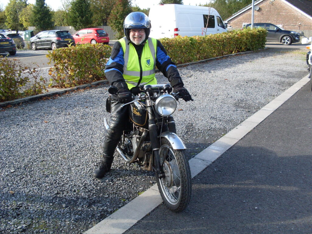 A portrait of a motorcycle, with a rider with a big grin, about to leave in the sunshine.