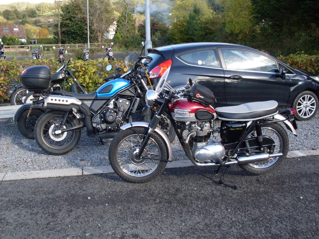 A red and grey Triumph twin and a more modern bright blue Honda each with a small handlebar-mounted screen
