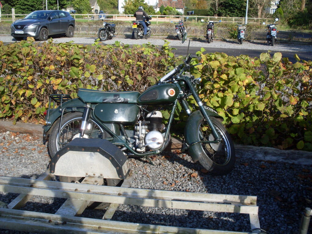 An elderly green Francis Barnet motorcycle has been parked behind the Honda and Triumph right next to the hedge and is leaning at an alarming angle