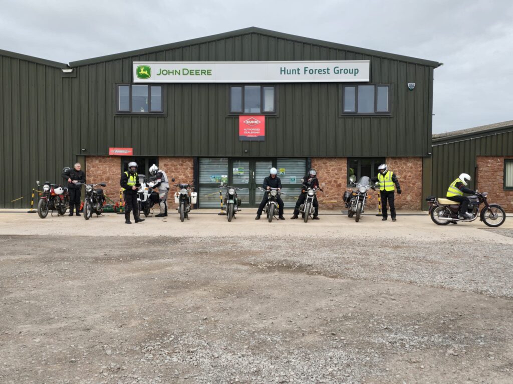 A line up of 9 motorcycles with riders variously standing and walking, in front of a modern green steel-clad building with a prominent 'John Deere' sign across it.