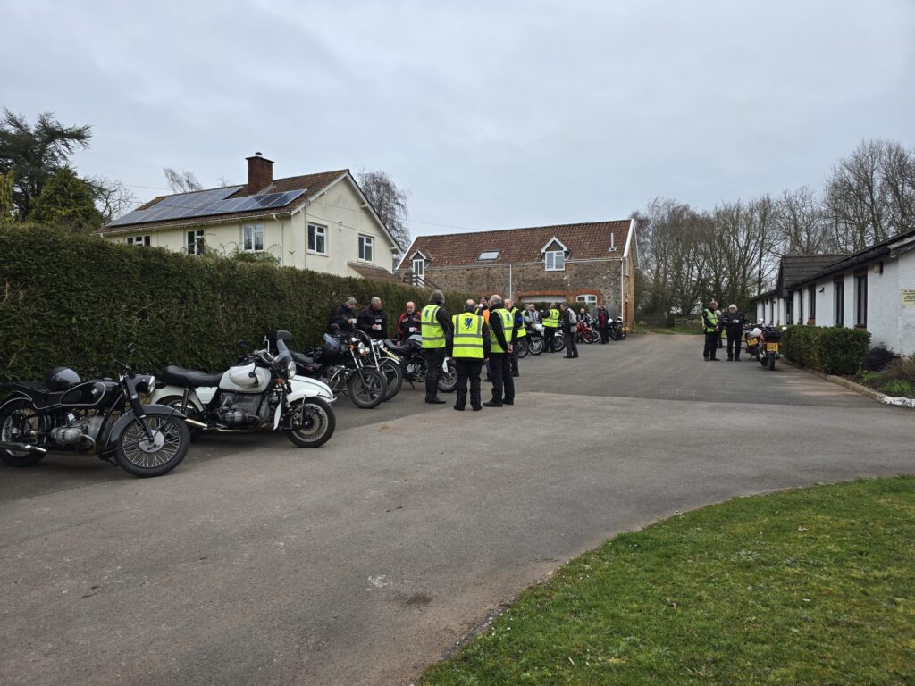 The line up of parked motorcycles in front of the hedge again but in this shot a large number of the chatting motorcyclists can be seen with mugs in their hands, or to their lips...!
