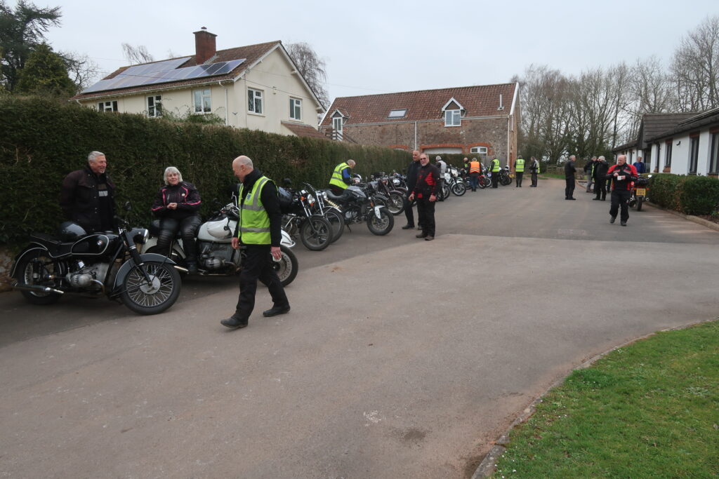 Parked motorcycles lined up in front of the hedge stretching back to the stone barn and other buildings in the background. Closest to the camera a black BMW solo with a male standing behind and a white BMW sidecar outfit with a female rider seated on it, both are grinning at another male walking past, obviously exchanging some banter!