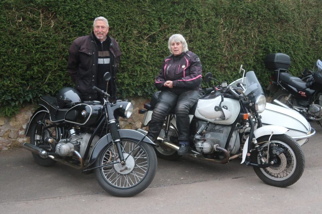 A black BMW motorcycle from the 1950s and a later (1970s) white BMW motrocycle and sidecar parked in front of a high hedge. The lady rider is perched on the saddle of the outfit, the male rider stands behind his earlier solo.