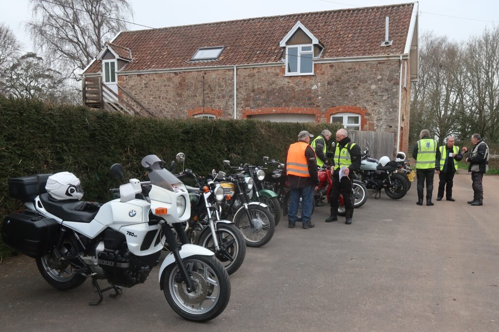 At teh start of te run, a line up of parked motorcycles in front of a high hedge with a converted stone barn in the background - and some small groups of people in riding gear, chatting amnong themselves.
