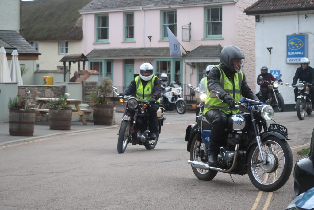 Several motorcyclists riding into a parking area, a village street with whitewashed houses and a pub forecourt with tubs of flowers behind them.