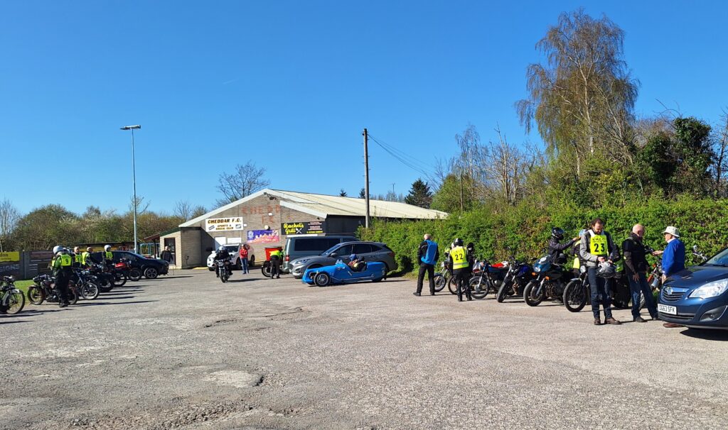 A different view of the car park with the football ground Clubhouse in the distance and cars and motorcycles parked on either side. The sky is bright blue and sunny, riders are wearing green bibs with numbers, some are walking towards their machines, some are standing next to them.