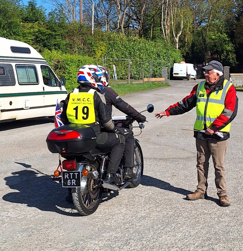 Rider and pillion (pillion with a green number 19 bib), from the back, wait next to the starter, who wears a green gilet and grey baseball cap and has an arm outstretched towards the bike, holding it at the ready...