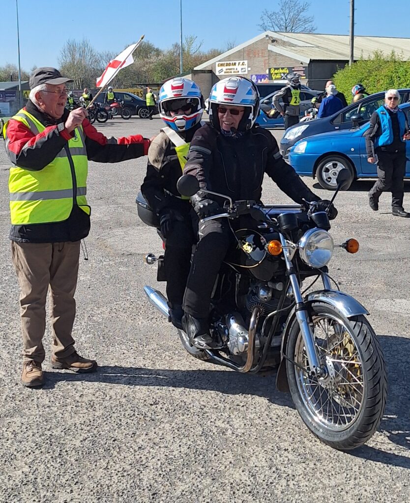 The same rider and pillion from the front, the starter now waving them away with a flag and the rider smiling cheerfully