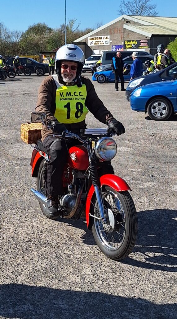 Against a backdrop of football ground buildings and parked vehicles a grinning motorcyclist (green bib no. 18) on a red bike approaches the camera