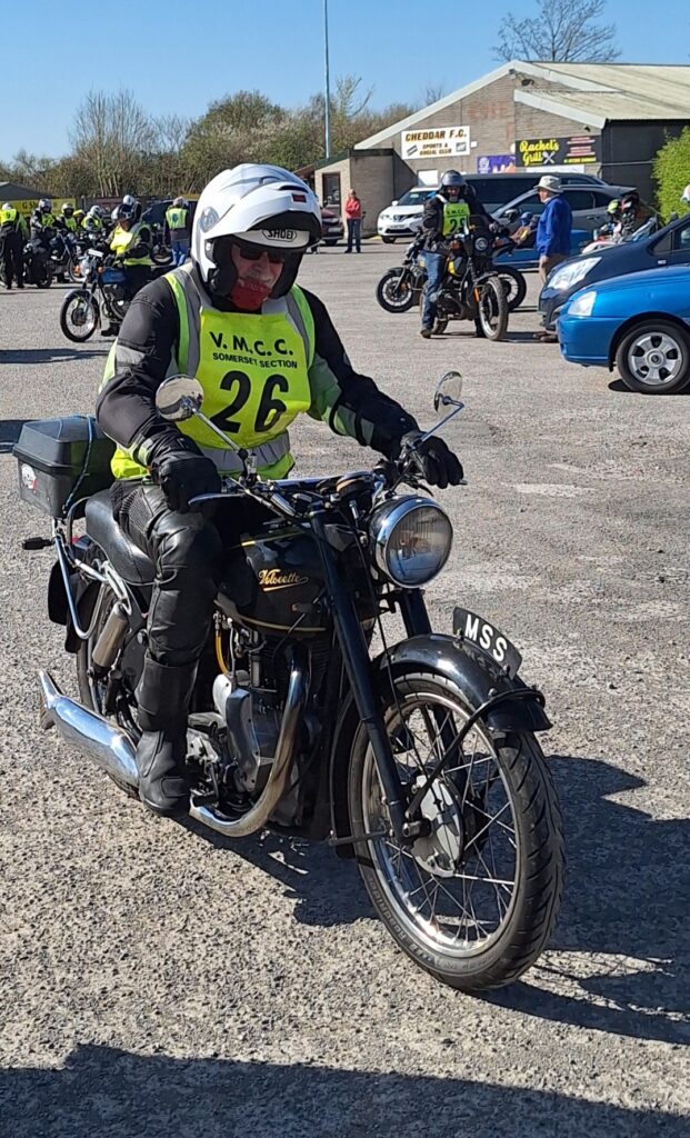 Against a backdrop of football ground buildings and parked vehicles a motorcyclist (green bib no. 26) advances, on a black single-cylinder bike.