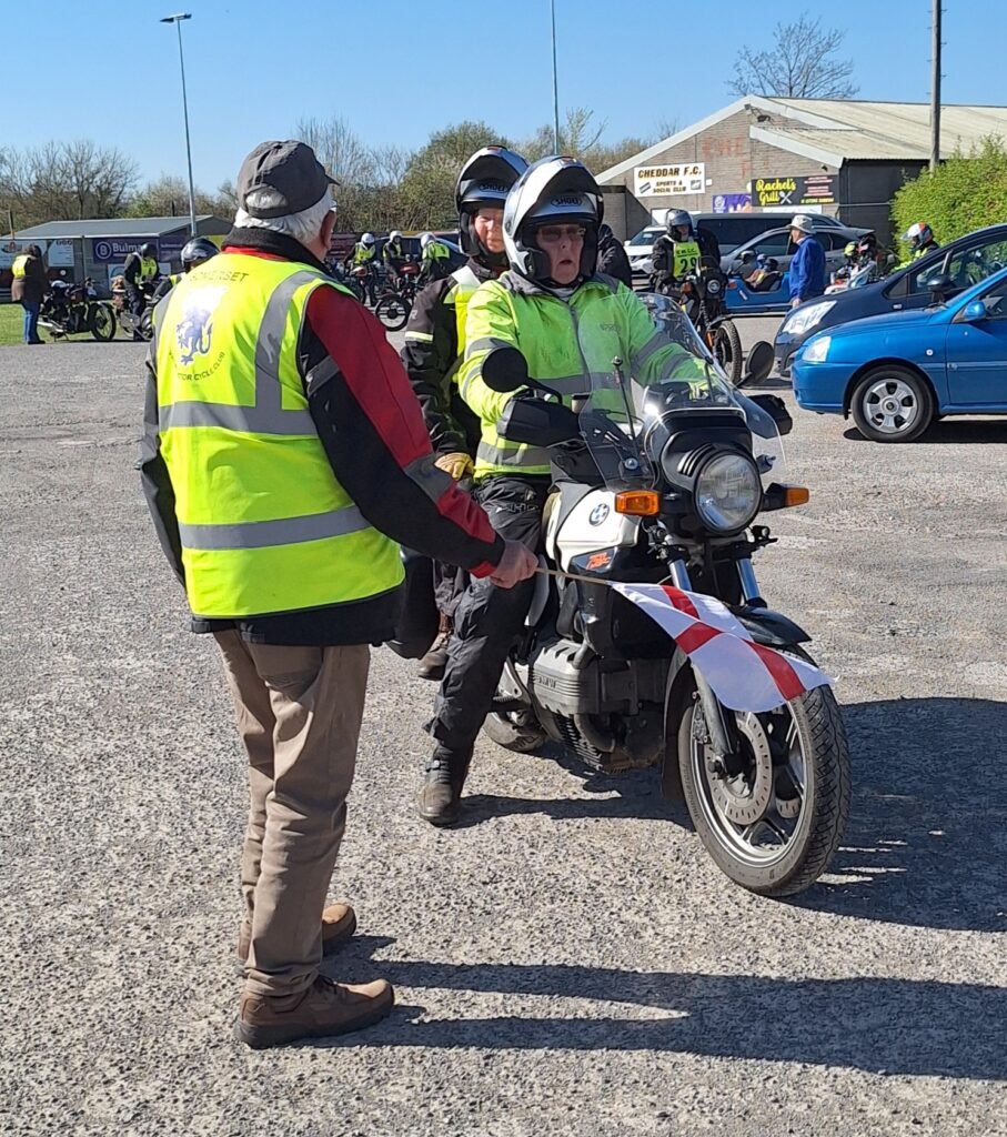 Against a backdrop of football ground buildings and parked vehicles a more modern BMW with rider and pillion draws up to the starter, who stands with back to camera and flag out horizontally to draw the bike to a stop.