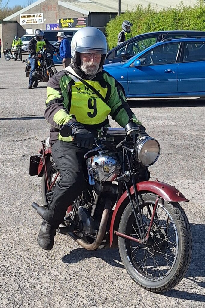 Against a backdrop of football ground buildings and parked vehicles a bearded motorcyclist (green bib no. 9) on an old red and chrome machine advances towards the camera