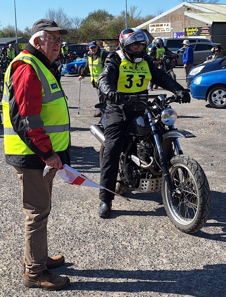 Against a backdrop of football ground buildings, parked vehicles and people a motorcyclist (green bib no. 33) waits next to the starer, both are looking pensively at something behind and to the right of the camera...