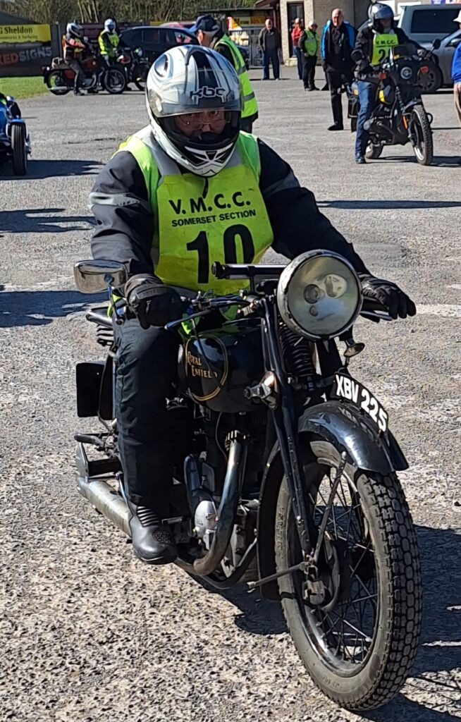 Against a backdrop of football ground buildings, parked vehicles and people a motorcyclist (green bib no. 10) on an old black machine with a big headlight advances towards the camera
