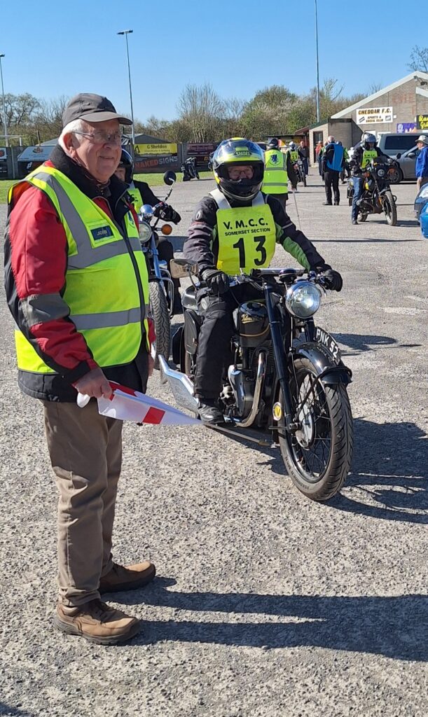 Against a backdrop of football ground buildings, parked vehicles and people a motorcyclist (green bib no. 13), on a black machine, waits next to the starter. Both are looking hard at something 'off screen' to the right of the camera