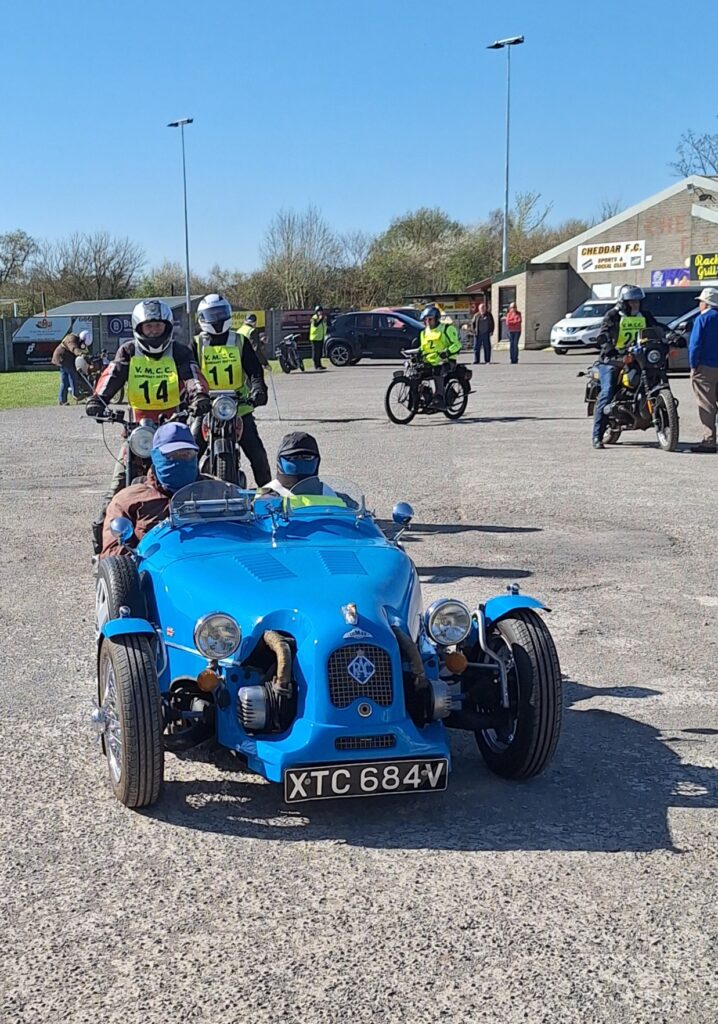 Against a backdrop of football ground buildings and parked vehicles two motorcyclists the bright blue Lomax three-wheeler advances towards the camera. Its two occupants have blue scarves pulled right up to their eyes and baseball caps pulled well down!