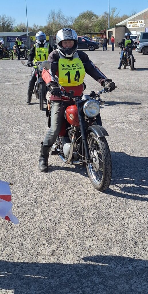 Against a backdrop of football ground buildings and parked vehicles two motorcyclists (green bib nos. 14 and 11) advance towards the camera, no 14 leading on a machine with a bright red petrol tank.