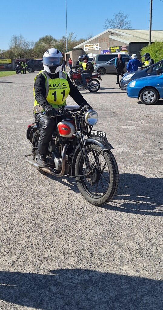 Against a backdrop of football ground buildings and parked vehicles a motorcyclist (green bib no. 11 ) advances towards the camera on a old single cylinder machine with a red and silver tank. The rider is sitting very upright looks directly at the camera through a full-face helmet that only reveals his dark sunglasses!