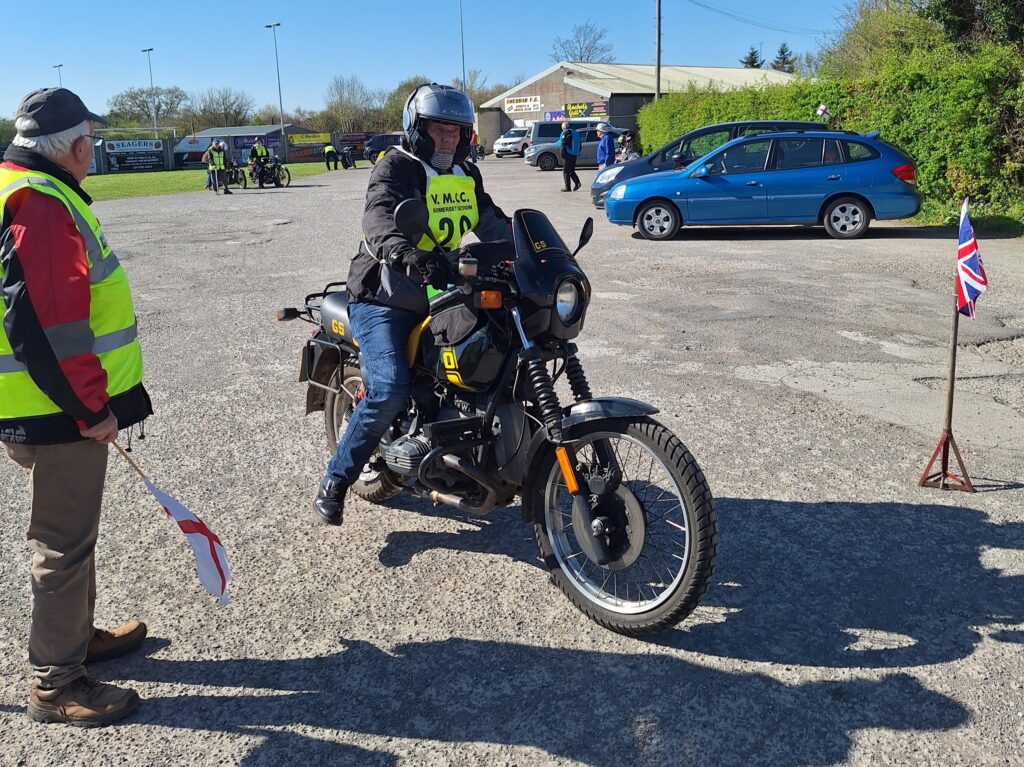 Against a backdrop of football ground buildings and parked vehicles a motorcyclist (green bib no. obscured) advances with towards the starter (who stands at left ) with a dangling foot for support