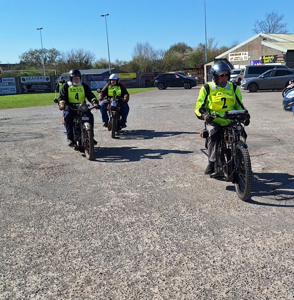Against a backdrop of football ground buildings and parked vehicles three motorcyclists (green bib nos. 2, 4 and 8) advance towards the camera