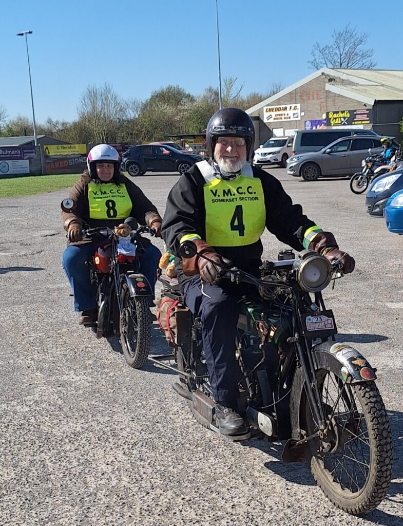Against a backdrop of football ground buildings and parked vehicles two motorcyclists (green bib nos. 4 and 8) advance towards the camera with broad grins!