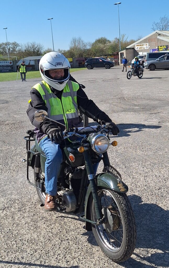 Against a backdrop of football ground buildings and parked vehicles a motorcyclist with blue jeans, white full-face helmet and green gilet (and apparently without a green bib) advances towards the camera on a dark green and black machine