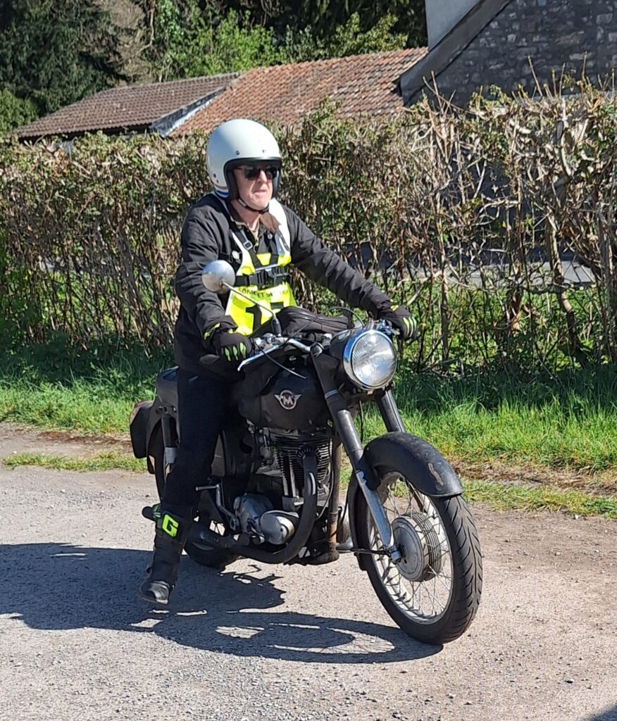 Against a backdrop of two tiled-roof buildings and a hedge a motorcyclist (green bib no. obscured) on a black machine waits with foot down.