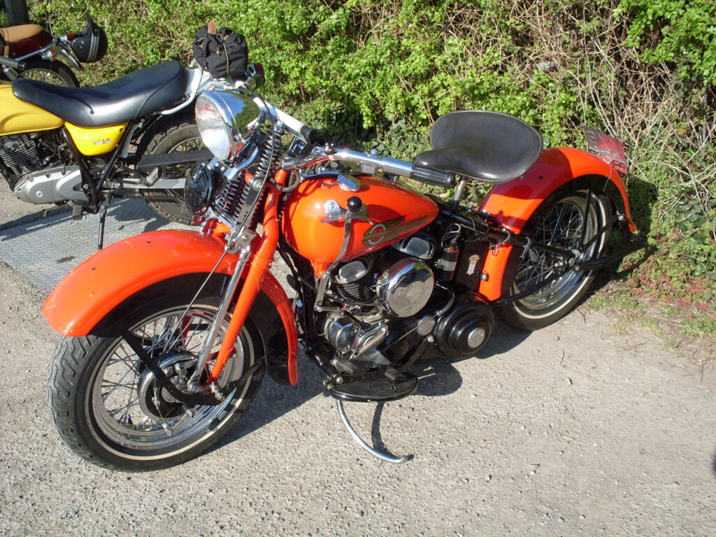 A red motorcycle with 'springer' forks, footboards and a big flat single seat parked on the gravel in front of a green hedge.
