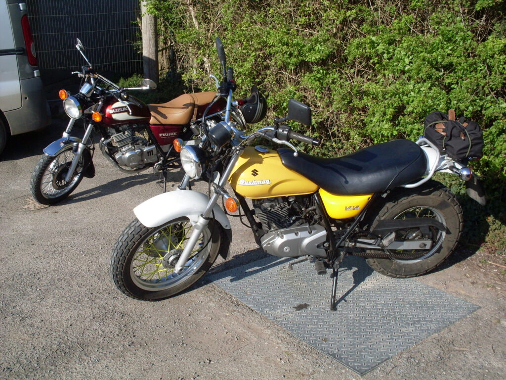 Two parked Suzuki motorcycles, a yellow 'Van Van Bushmaster' and behind an older red TU250X with a big light brown seat.