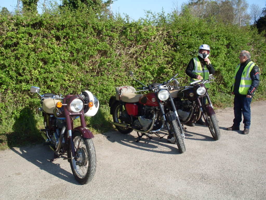 Three similar-looking red motorcycles parked in front of a green hedge with two of the riders (one taking his helmet off) talking behind them