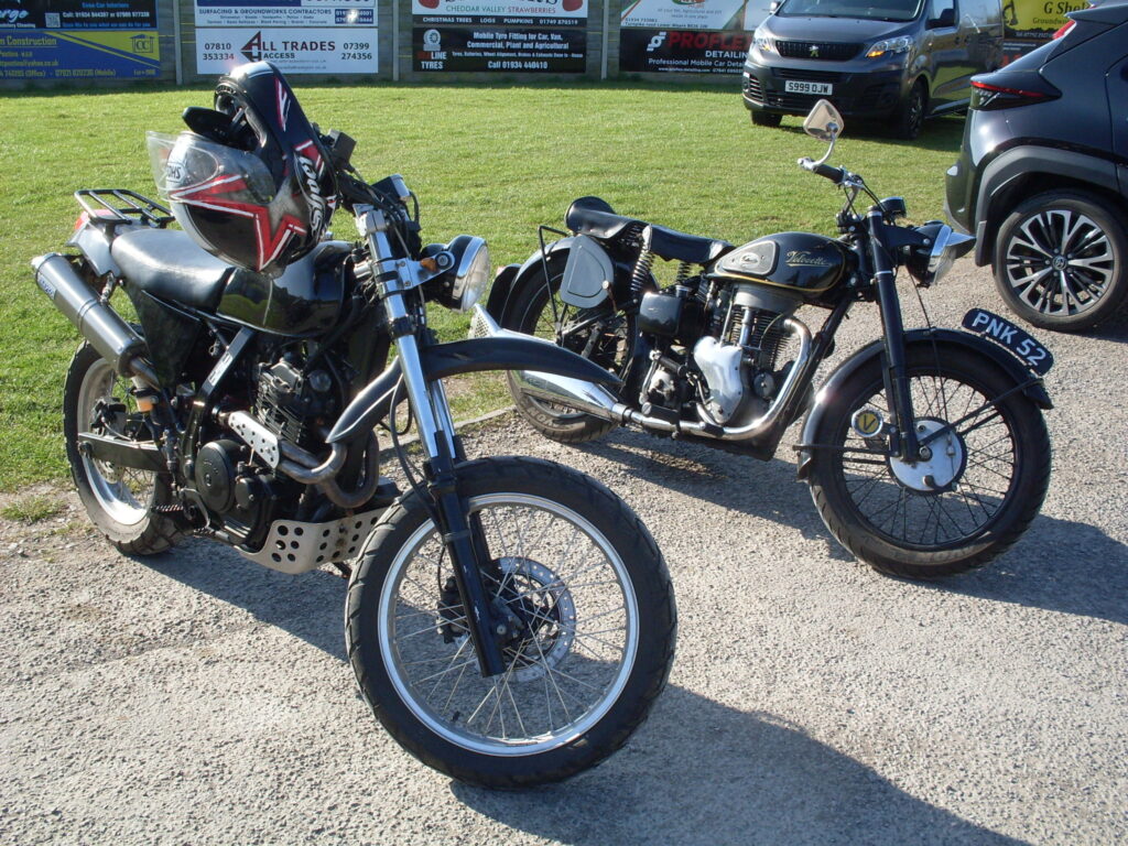 Two black motorcycles parked in the sun, the green grass of the playing ground behind them (and some advertising hoardings!)