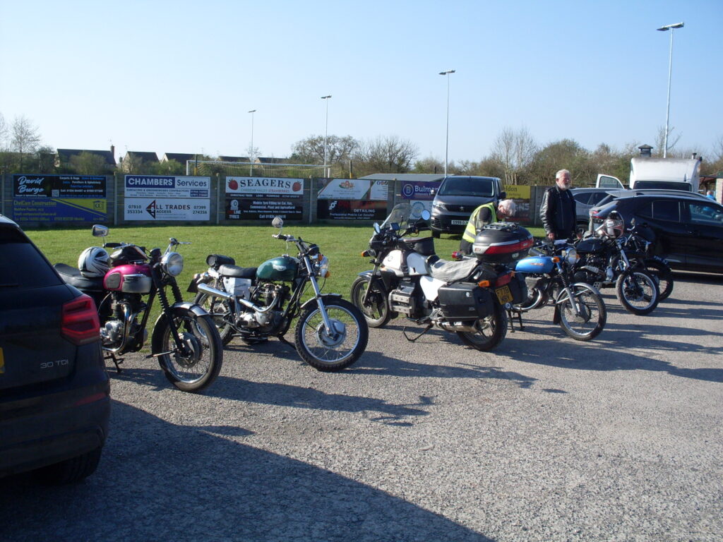 A short line up of varied motorbikes parked between two cars, a couple of riders, green grass (and advertising hoardings!) in the background, in bright sunshine and under a beautiful blue sky.