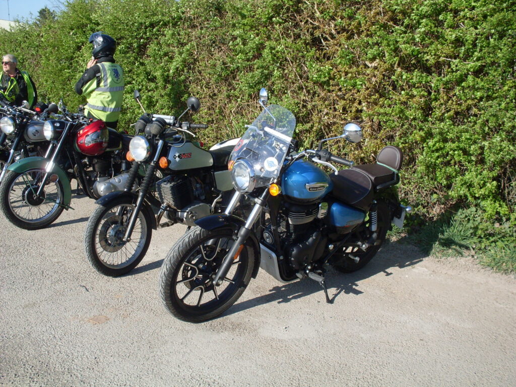 Three bikes of quite different ages parked up with a green hedge in the background - in the foreground a modern Royal Enfield with a blue tank and a tall flyscreen.