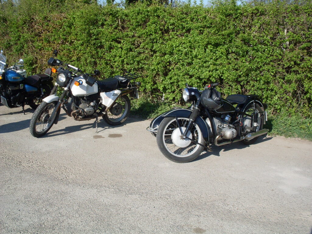Two BMW 'boxer' twin motorbikes parked in the sun in front of a green hedge, an older black one with a sidecar on the continental side and th eold-design engine and a much later white solo with the newer design engine.