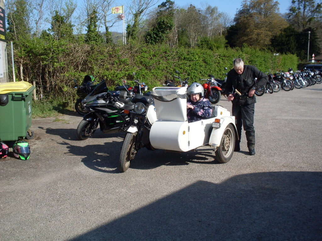 A white BMW sidecar outfit parked in front of a line of motorcycles, the lady passenger has just got in (the front of the single-seater open sidecar is still raised up to allow access); the black-suited male rider stands behind packing his pockets!