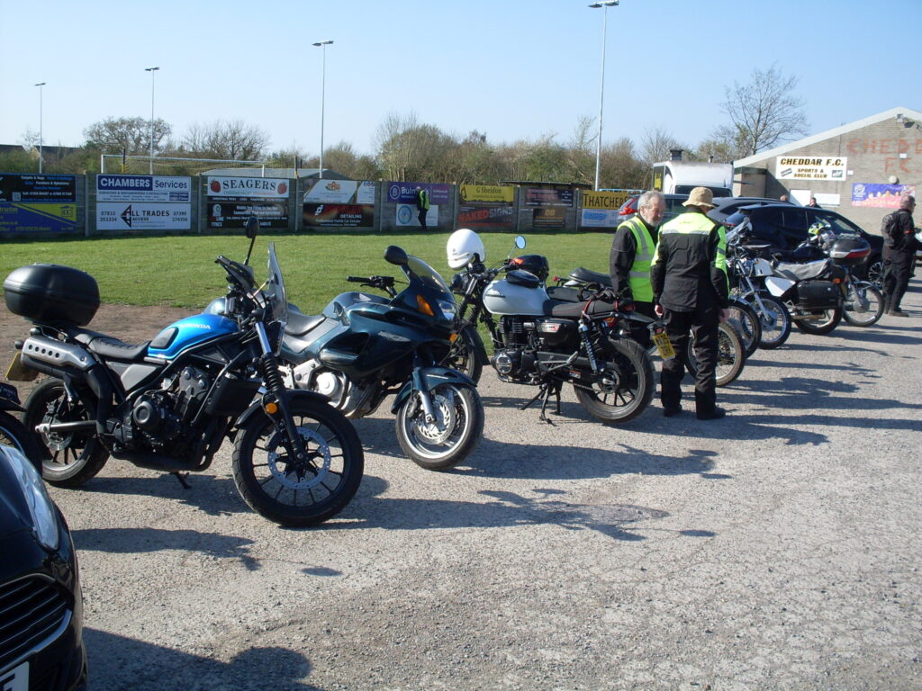 Several motorcycles parked in a line in front of the grass with hoardings, the football ground floodlight posts and clubhouse behind - two gents in riding gear (one in a sun hat and one with a green bib) stand talking halfway down the line...
