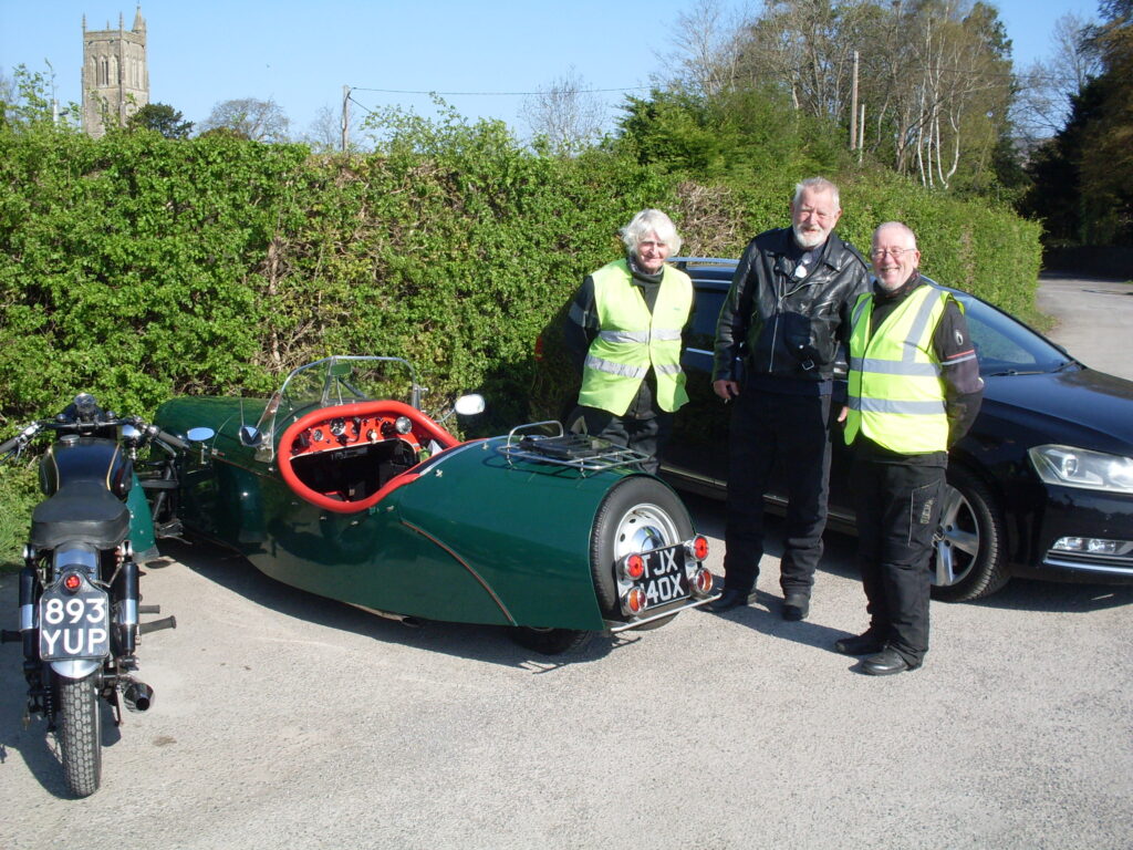 A rear view of a motorcycle, a three-quarter rear view of a green three-wheeler, three gentlemen in black riding gear (but two with green bibs) face the camera with big grins on their faces, in the sun.