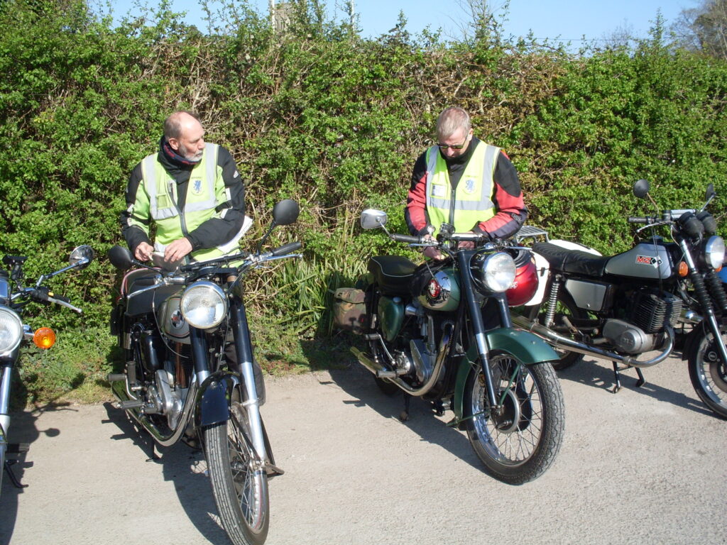 A front-on view of four parked motorbikes but the two central ones are prominent and their two riders stand behind them with their attention on preparing their machines.