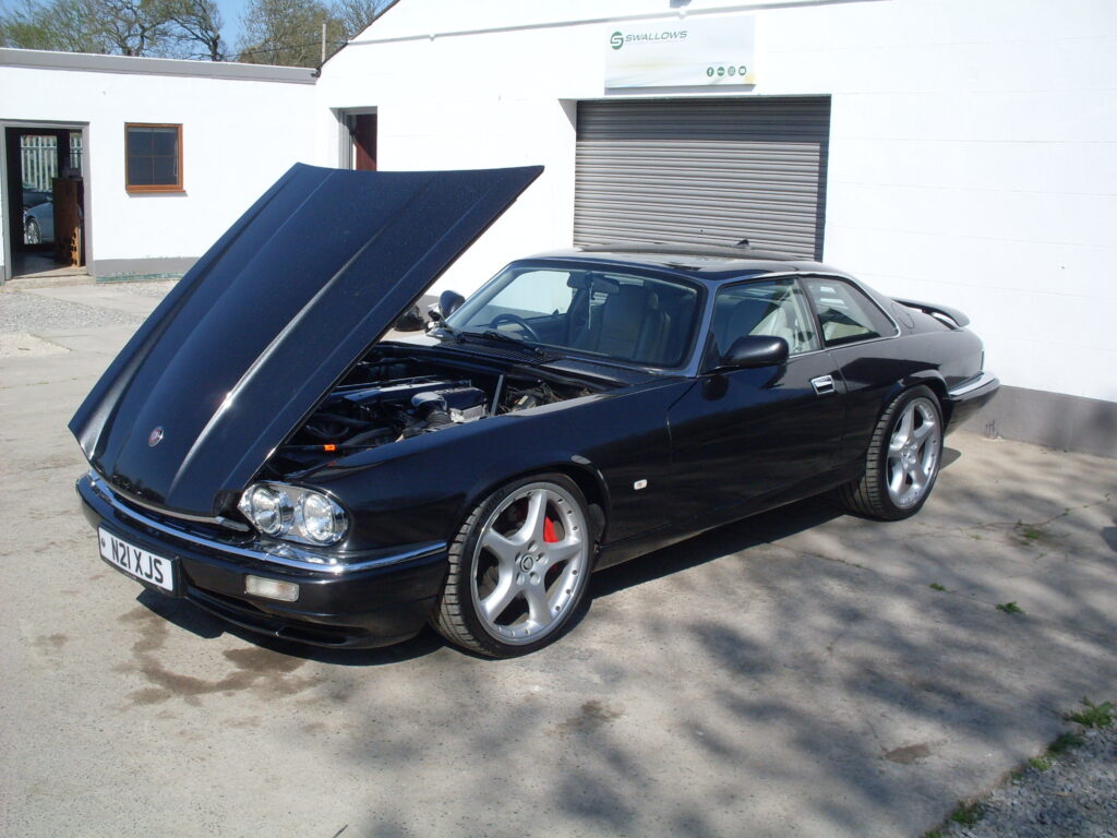 A black Jaguar XJS with tinted glass and 5-spoke alloy wheels is parked in front of some low white buildings with its bonnet open.