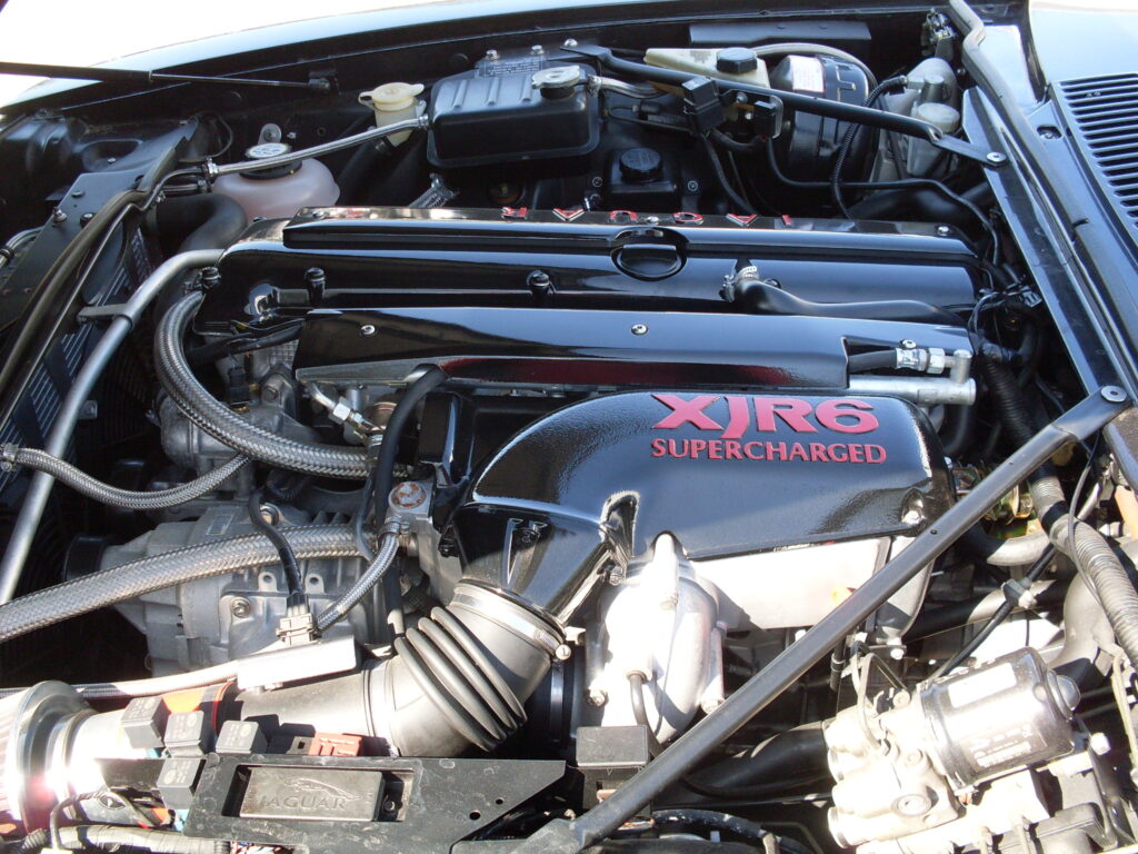 The engine bay of a car crammed with shiny black engine parts and clean aluminium fittings ; in the foreground a shiny black item with a big tubular duct attached and on its top the words (in red) 'XJR6 supercharged'