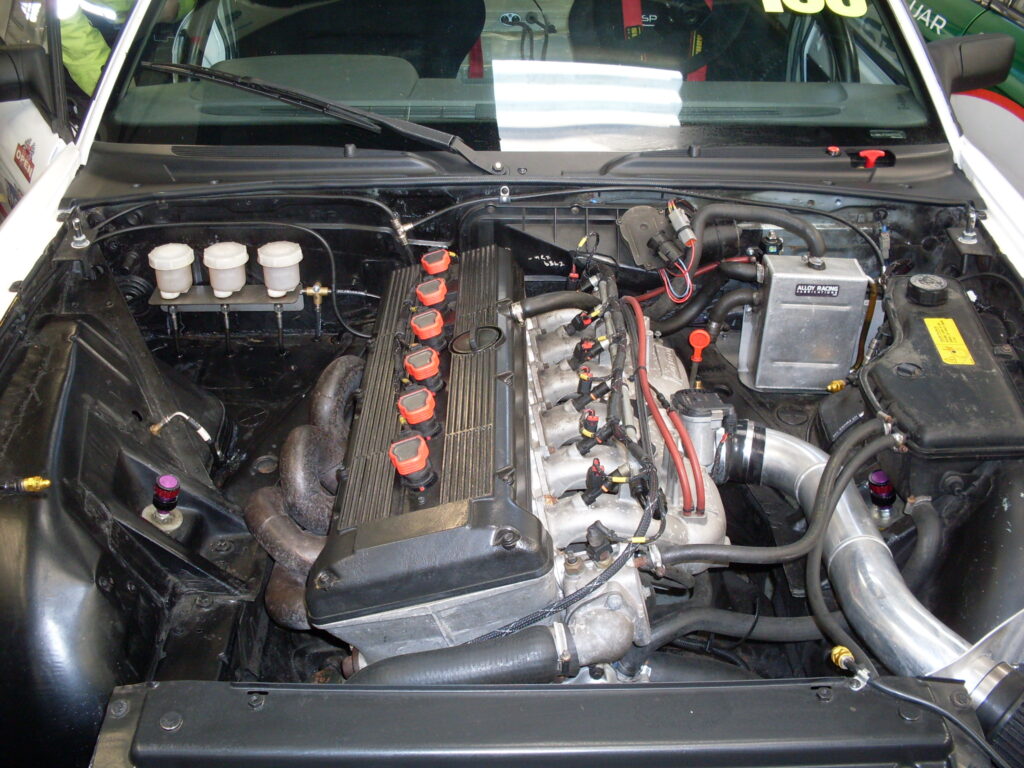 Front view of windscreen and black engine bay of a (white) car (the bonnet has been taken off) showing the black ribbed cam-cover, prominent aluminium inlet pipework and red electrical accessories of a big six-cylinder engine.