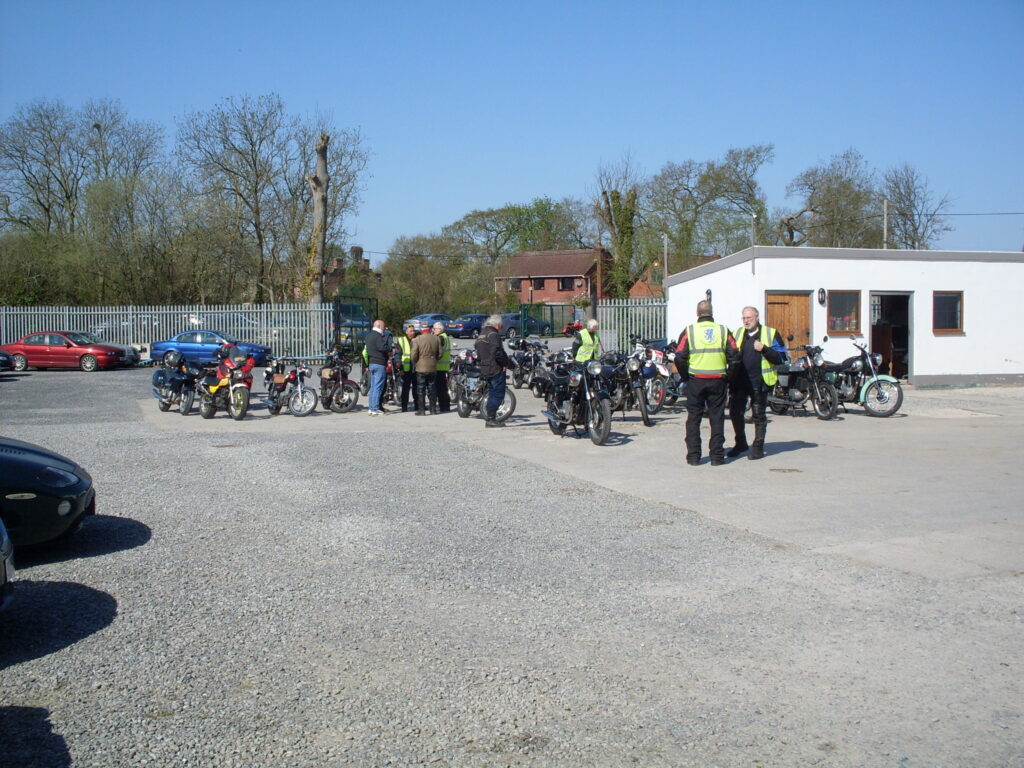 A group of motorbikes parked in the middle of a car park, with a low white office building behind at right and a background of a fence, tall trees and a few cars . Several people stand in small groups, in the sun, under a blue sky.