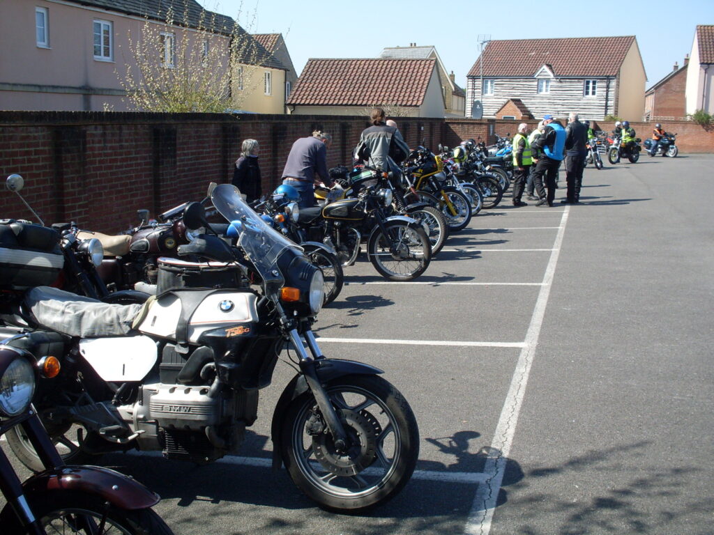 A big car park, tarmacked, parking bays delineated with white lines in front of a tall brick wall separating it from the houses behind - in front of the wall (stretching into the distance) is a line of parked motorbikes with riders standing, attending to the machines or talking between themselves.
