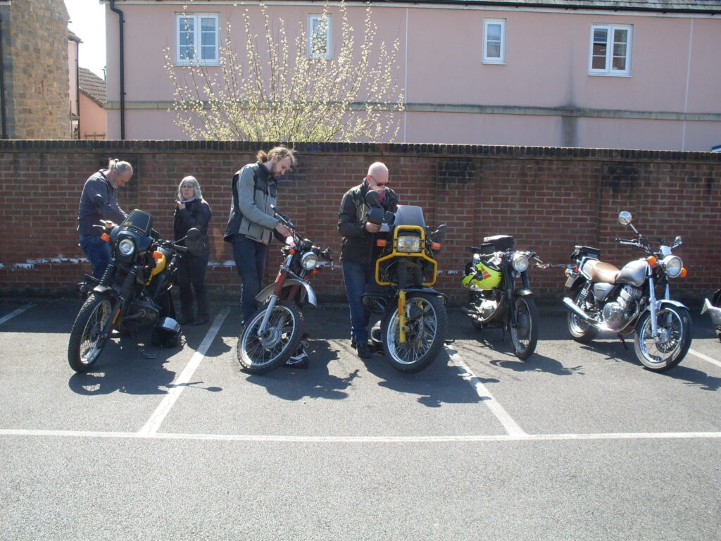 Several bikes in the parking bays in front of the brick wall (with pink-washed houses directly behind). Four riders 'adjust' their machines!