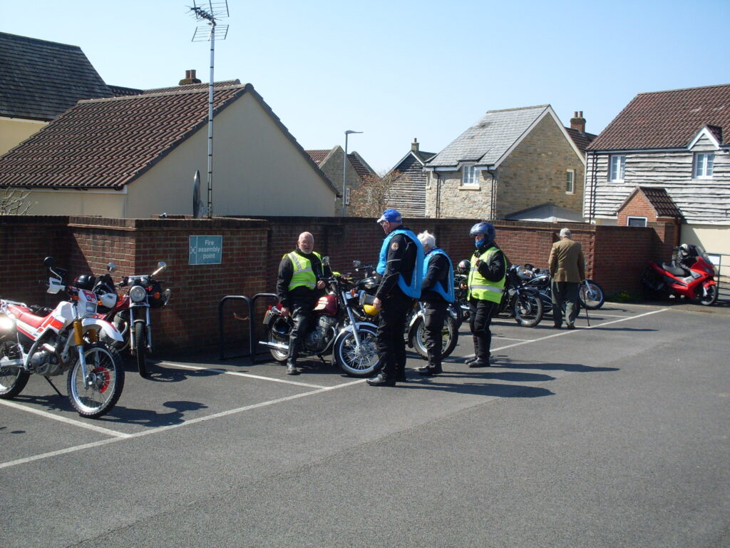 At the far end of the carpark, with houses behind the brick wall, several parked machines and riders - a small group in the centre in front of some bikes and on the wall behind them a big notice 'Fire Assembly Point' .